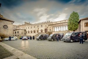 Furgoni e persone in piazza con edificio storico.
