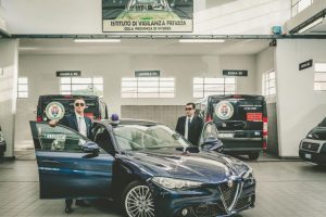 Due uomini in abiti formali accanto ad auto di sicurezza.